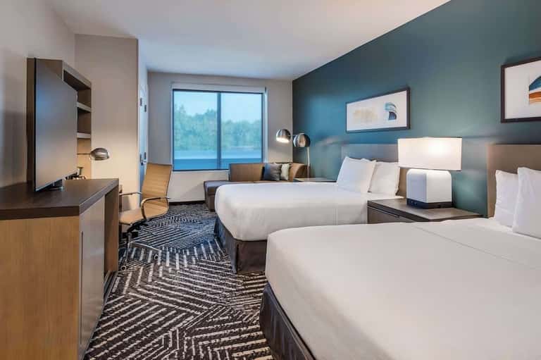 Double queen hotel room with teal walls, waterfront view windows, work desk, and contemporary furnishings