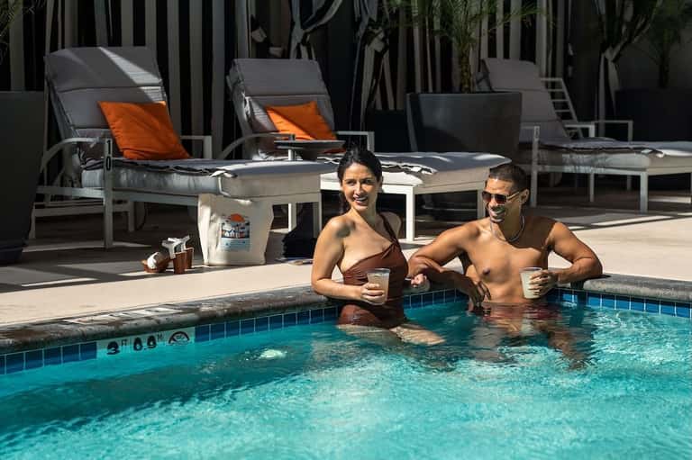 Two guests enjoying drinks in resort pool with loungers and striped umbrellas poolside