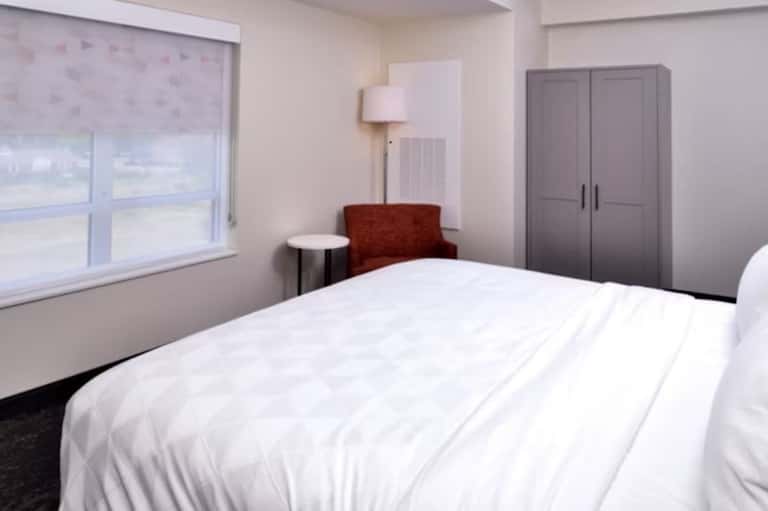 Modern hotel bedroom with queen bed, window, chair, lamp, and closet in neutral tones