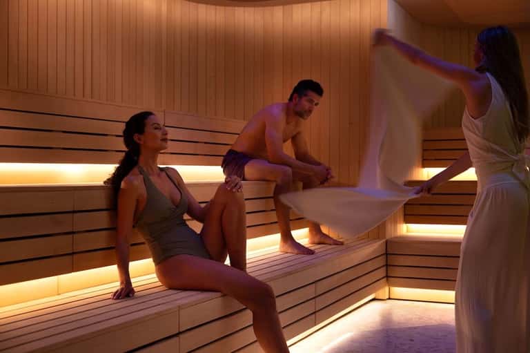 Contemporary sauna interior with wooden benches, warm ambient lighting, and three people relaxing