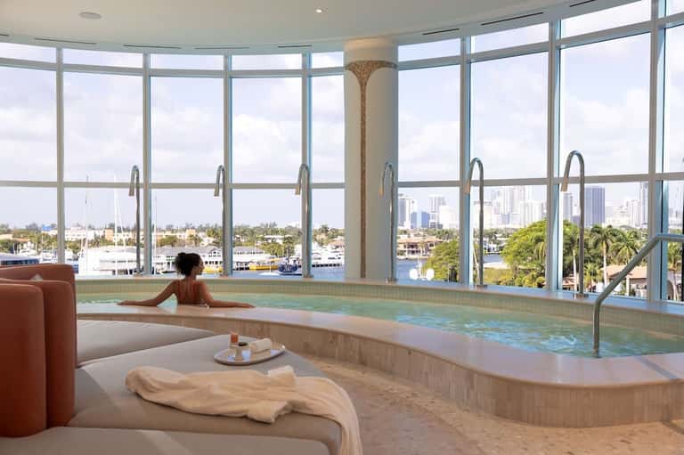 Luxury indoor pool with floor-to-ceiling windows overlooking waterfront city skyline, with lounge seating and towels