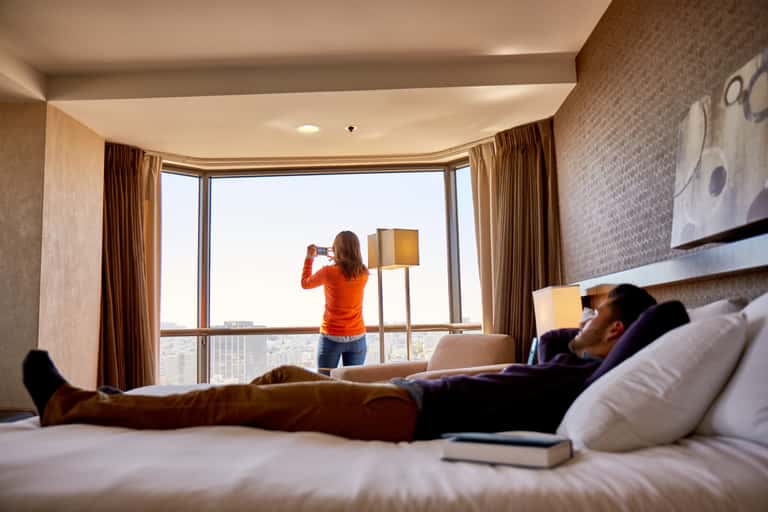 Hotel room with guests relaxing on bed while woman enjoys city view from floor-to-ceiling window