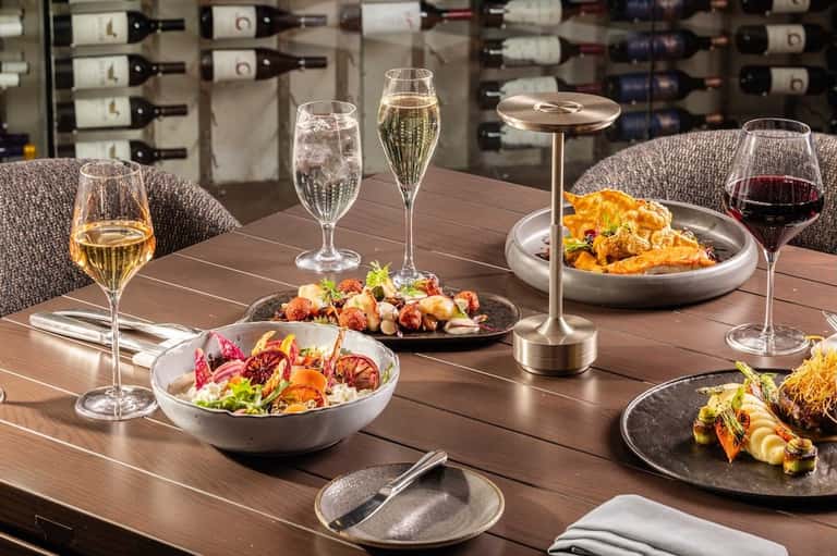 Fine dining table with gourmet dishes, wine glasses, and champagne, wine storage visible in background