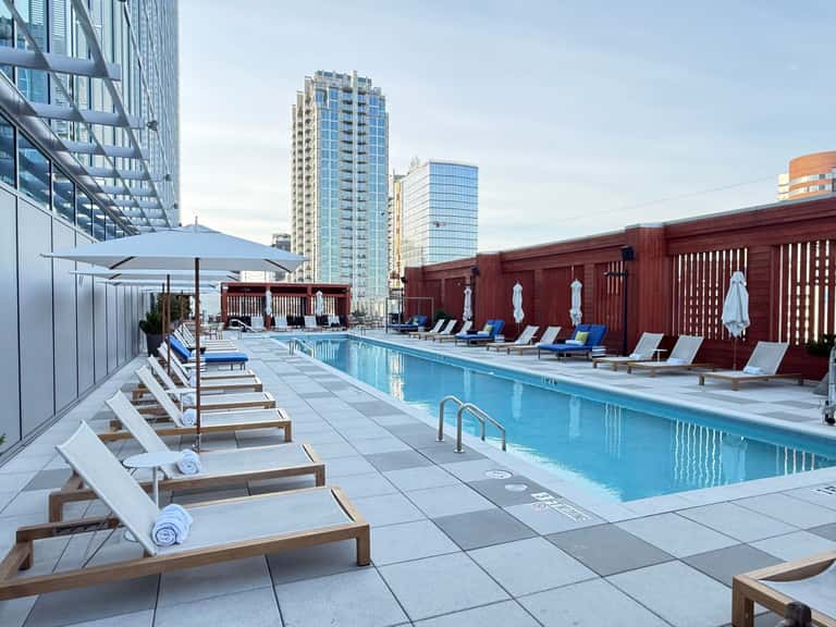 Rooftop pool deck at urban hotel with blue loungers, umbrellas, red wooden fence, and city skyline