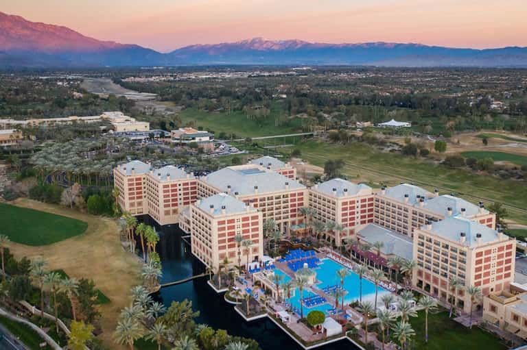 Aerial view of large pink resort with blue pool, palm trees, and snow-capped mountains in background at sunset