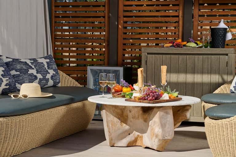 Outdoor patio with woven lounge chairs, round wooden table with fresh fruit and flowers, and wooden privacy screens