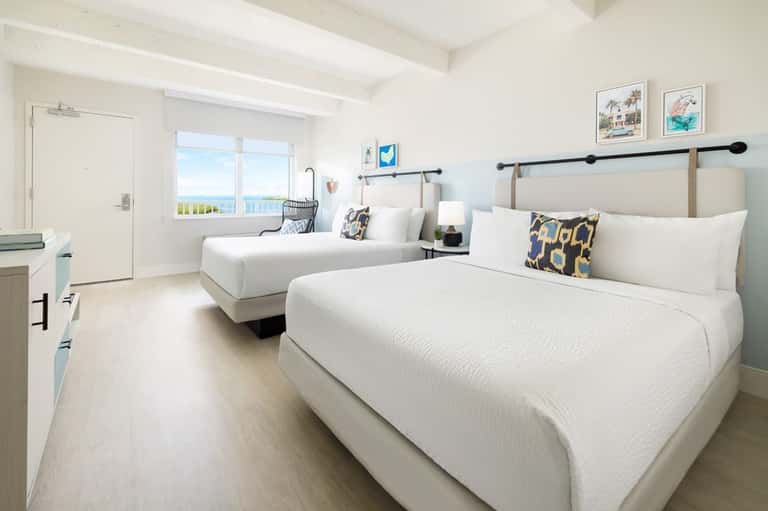 Bright hotel bedroom with two beds, ocean view balcony, and coastal decor