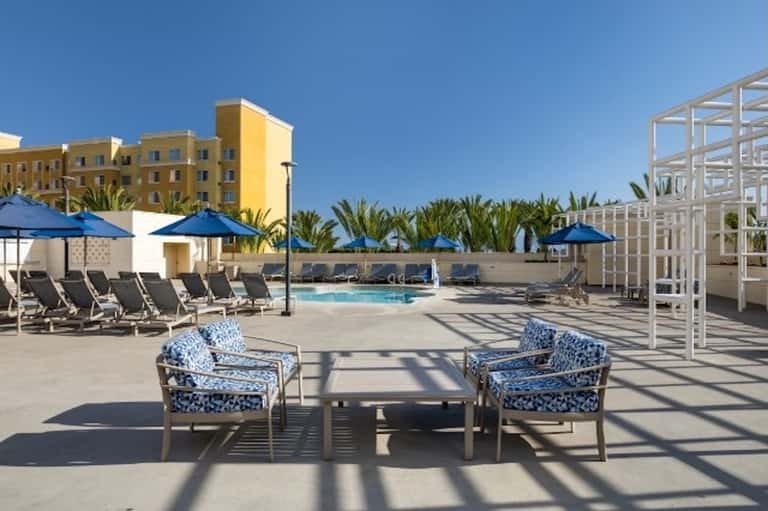 Resort pool deck with blue umbrellas, lounge chairs, white pergola, and palm trees