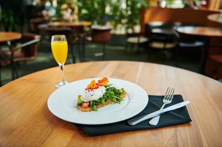 Plated brunch dish with avocado toast, egg, and citrus juice on wooden table in bright restaurant