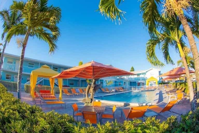 Resort pool with orange and yellow umbrellas, palm trees, turquoise water, and blue buildings.