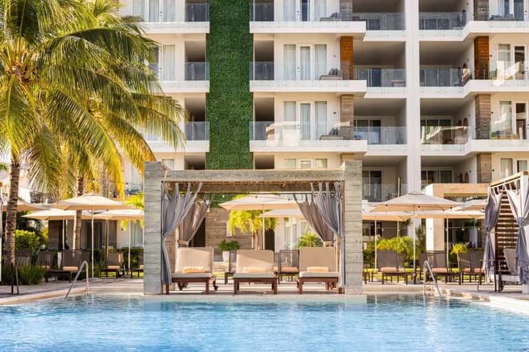 Modern resort pool area with white umbrellas, wooden pergolas, multi-story building, and palm trees