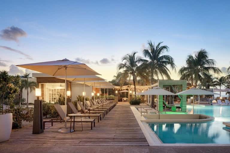 Resort pool deck at dusk with lounge chairs, large umbrellas, modern architecture, and palm trees