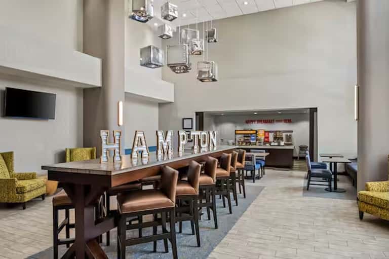 Hotel lobby and breakfast area with high ceilings, HAMPTON sign, bar seating, and modern décor