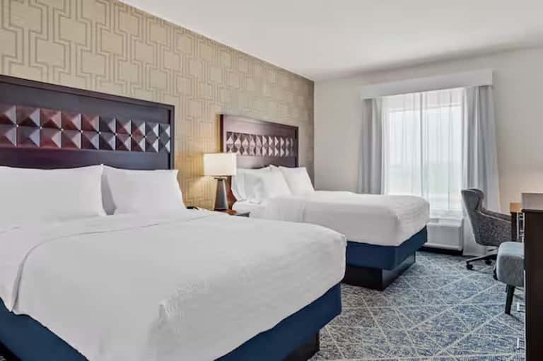 Hotel double queen room with white bedding, blue bed base, patterned wallpaper, and balcony access
