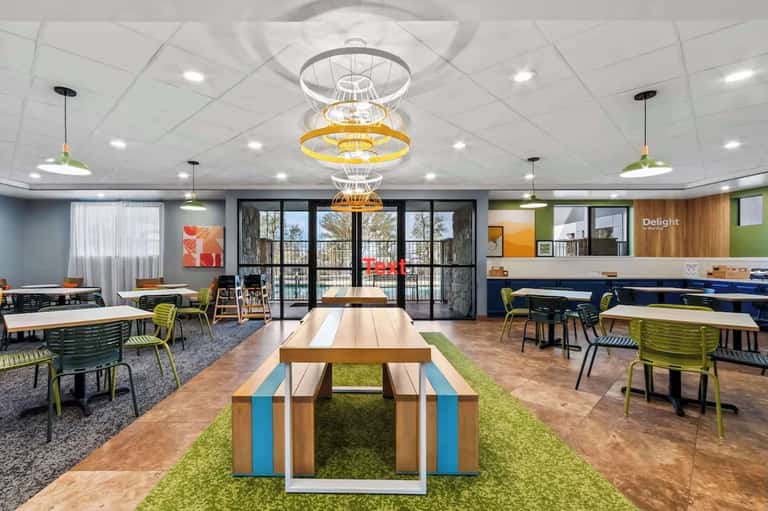 Bright communal workspace with long wooden tables, colorful chairs, large windows, and hanging pendant lights overlooking balcony