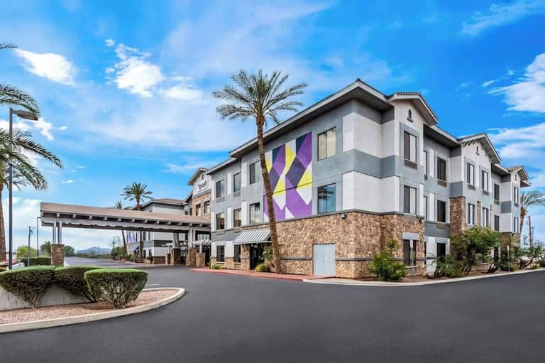 Hotel side entrance with covered portico, stone base, white upper floors, purple accent signage, and desert landscaping