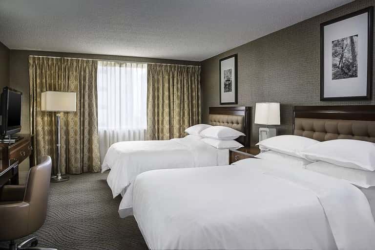Hotel double room with two queen beds, patterned curtains, desk, and traditional furnishings in neutral tones