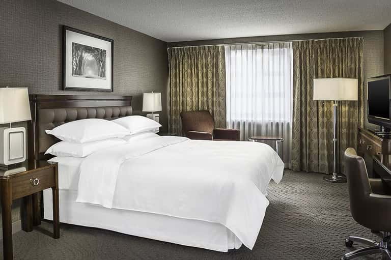 Upscale hotel suite with king bed, sitting area, elegant curtains, and taupe textured wallpaper throughout