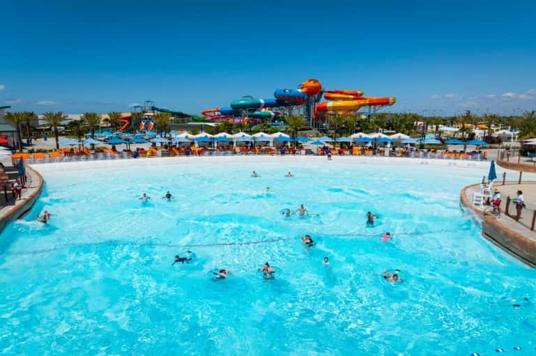Large wave pool with swimmers and colorful water slides in the background at a resort
