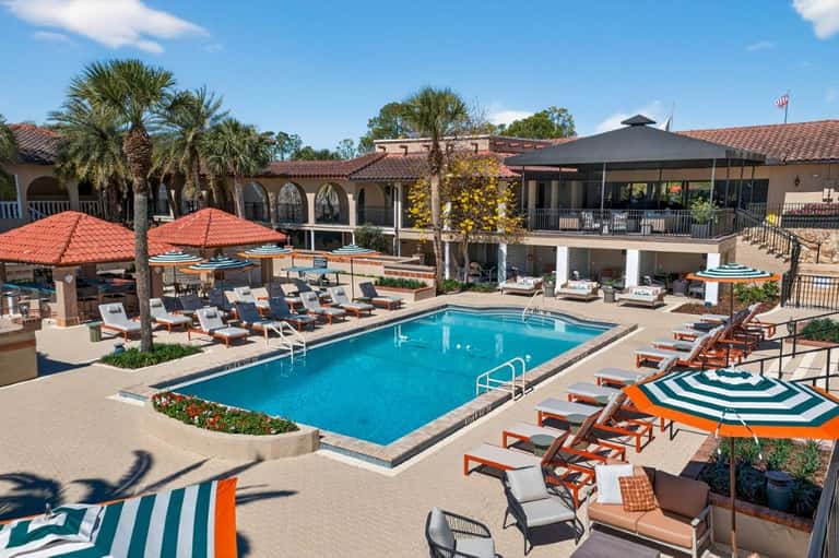 Hotel pool deck with colorful umbrellas, lounge chairs, and Spanish-style architecture with palm trees