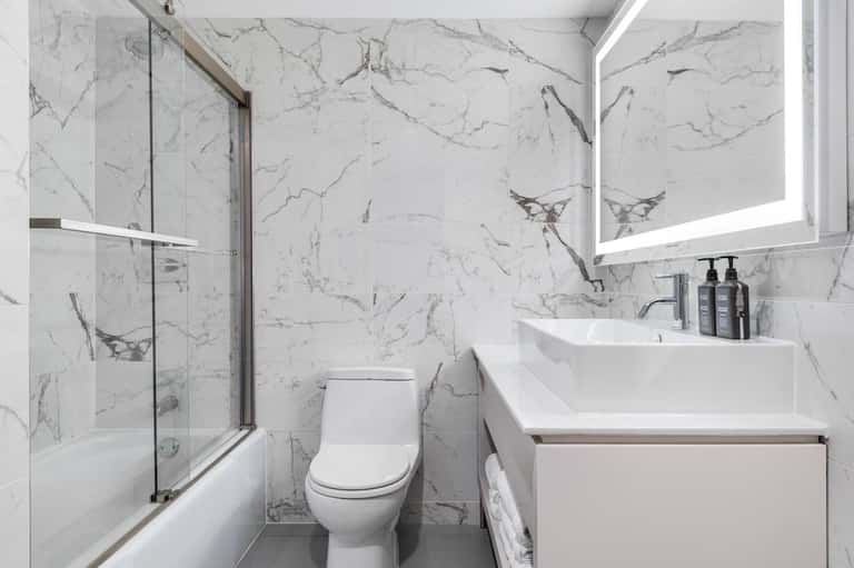 Modern bathroom with marble-tiled walls, glass-enclosed shower, white fixtures, and LED mirror