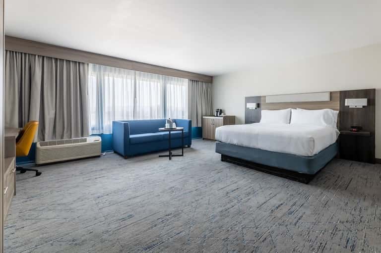 Spacious hotel suite with king bed, blue sofa seating area, floor-to-ceiling windows, and modern wood accents