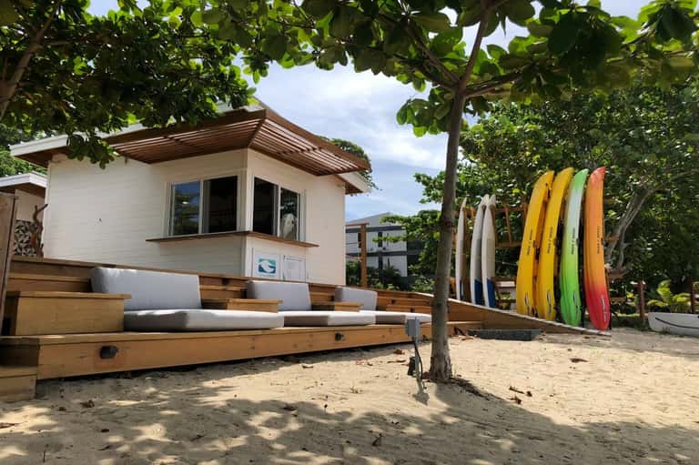 Beach shack with white walls and colorful surfboards leaning against trees on sandy ground