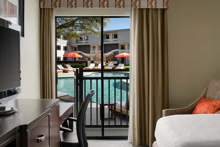 Hotel room with desk, bed, and balcony overlooking pool area with umbrellas and lounge chairs