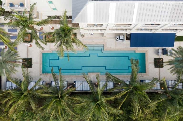Aerial view of a rectangular turquoise swimming pool surrounded by white buildings and palm trees