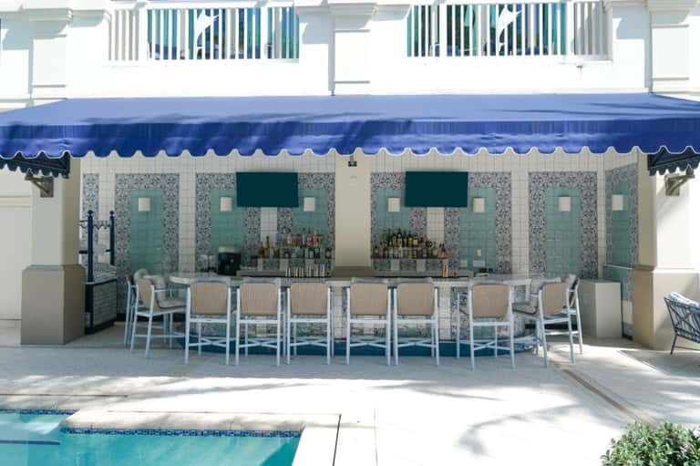 Poolside bar with turquoise tile accents, white bar seating, and blue awning overlooking the pool