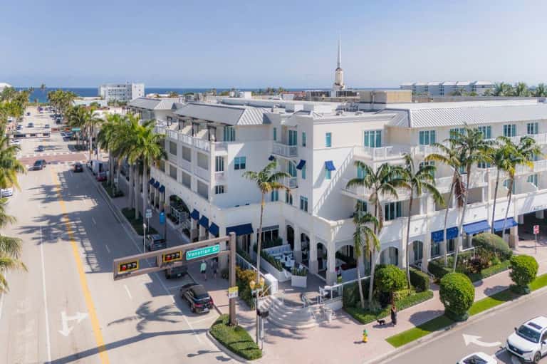 White beachfront resort buildings with blue accents and palm trees lining a street near the ocean