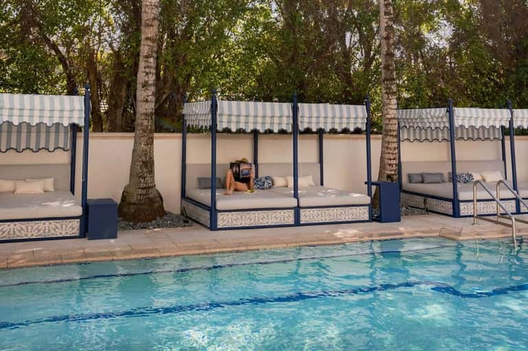 Luxury cabana beds with white striped canopies along a resort pool surrounded by mature trees
