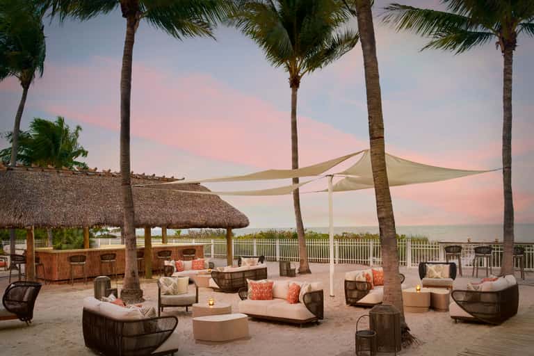 Beachfront lounge area with woven seating, fire pit, and sail shade at sunset with palms