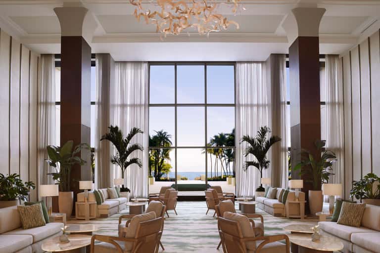 Luxurious lobby with tall windows, palm plants, cream furnishings, and ocean views