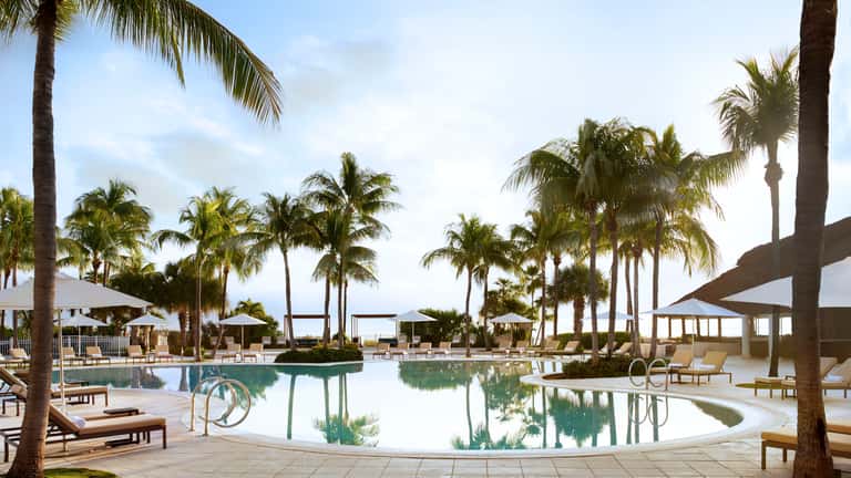 Large lagoon-style pool surrounded by white umbrellas, palm trees, and modern resort structures