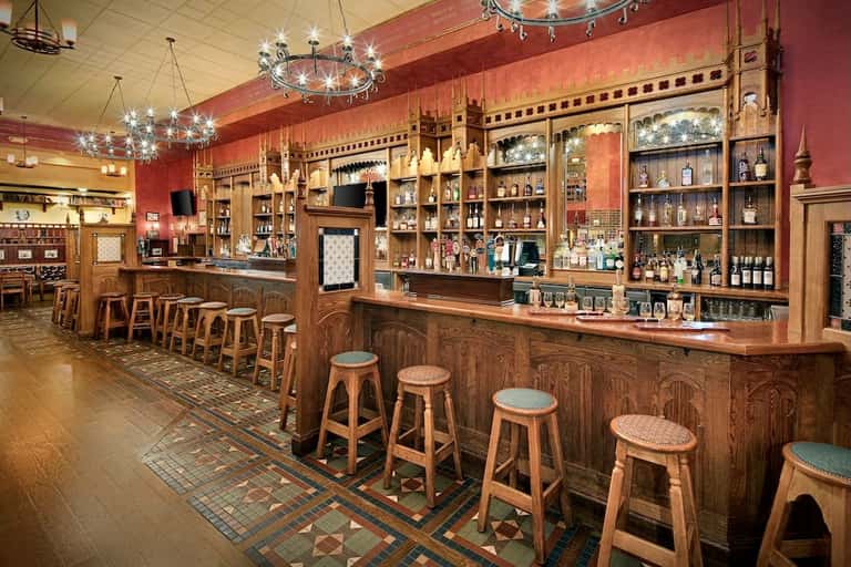 Upscale bar interior with ornate wood cabinetry, red walls, decorative chandeliers, and extensive liquor bottle display