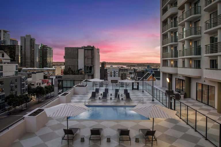 Rooftop pool at dusk with pink sunset sky, striped umbrellas, and city skyline backdrop