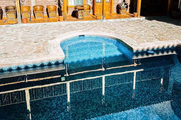 Kidney-shaped pool with blue mosaic tiles, stone decking, and wooden chairs beside building
