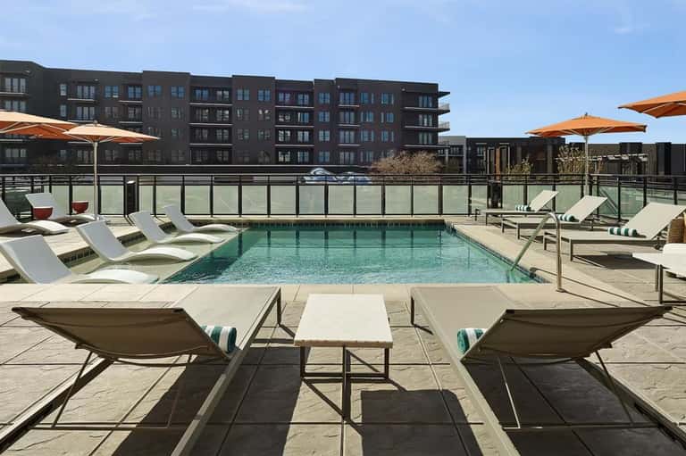 Rooftop pool deck with loungers, orange umbrellas, and modern dark apartment buildings in background