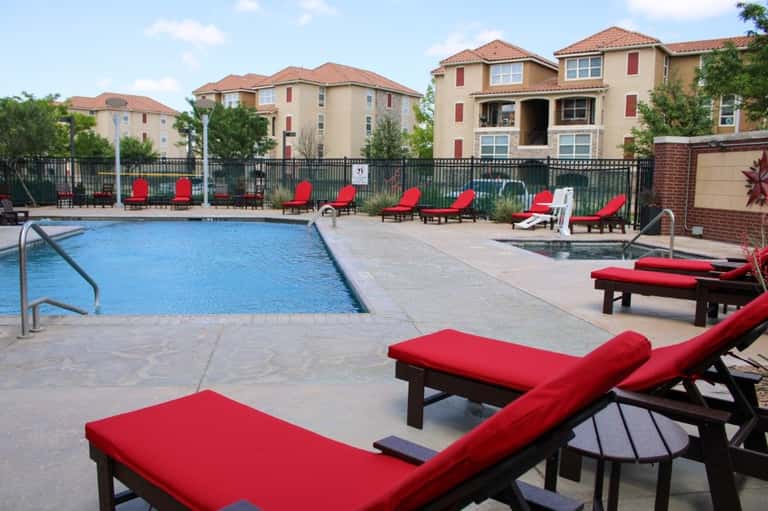 Resort pool with blue water, red lounge chairs, and tan apartment buildings with terracotta roofs in background
