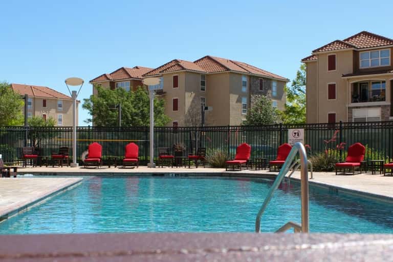 Swimming pool fronted by red cushioned lounge chairs and black fence with tan residential buildings behind
