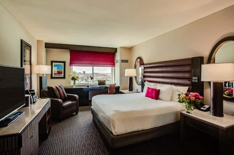 Hotel bedroom with king bed, burgundy headboard, burgundy window shade, dark wood furniture, and city view