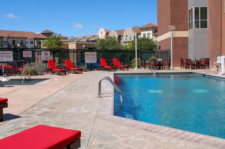 Outdoor swimming pool with blue water, red lounge chairs, brick residential building, and black perimeter fence