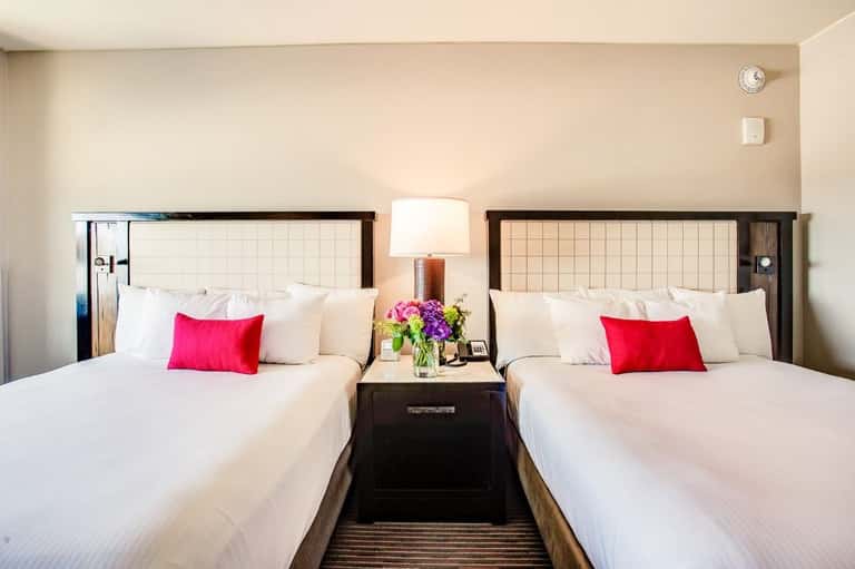 Hotel room with two queen beds, white linens, red accent pillows, and burgundy floral arrangement between beds