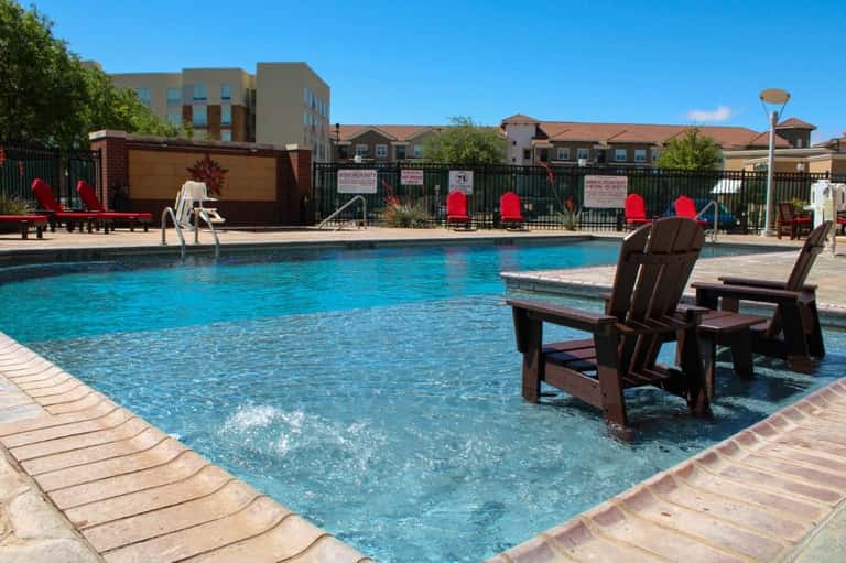 Resort pool with lounge chairs partially submerged in water and red cushioned chairs poolside near tan buildings