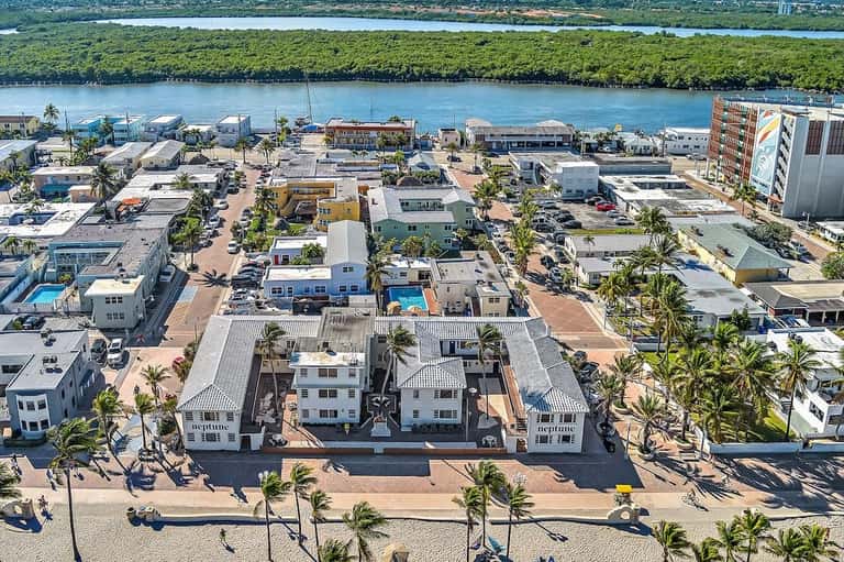 Aerial view of beachfront resort with white buildings, pool, palm trees, and river in background