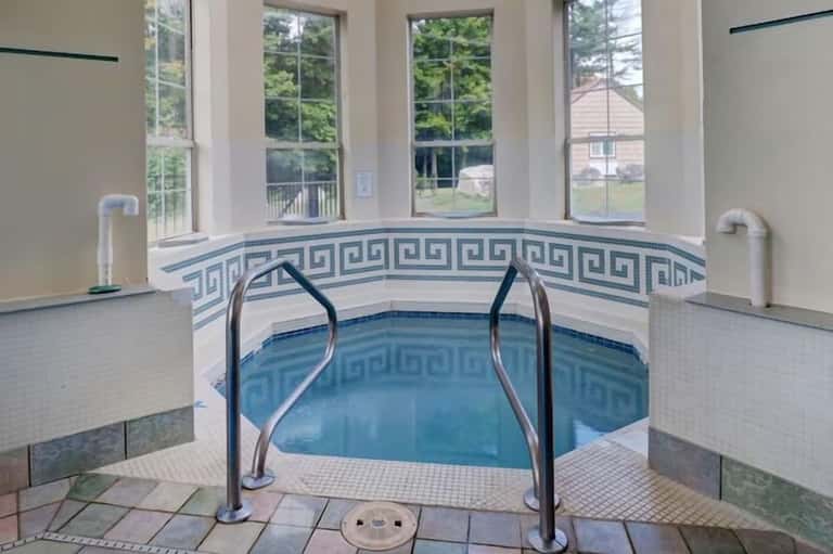 Indoor hot tub with Greek key tile pattern, metal handrails, large windows overlooking green landscape