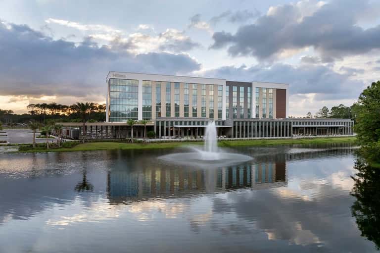 Contemporary Hilton hotel exterior reflecting in calm water with fountain, surrounded by green landscape at dusk