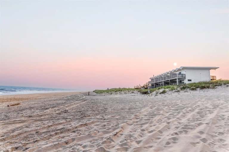 Beachfront house at dusk with pink sky, moon visible, sandy beach in foreground