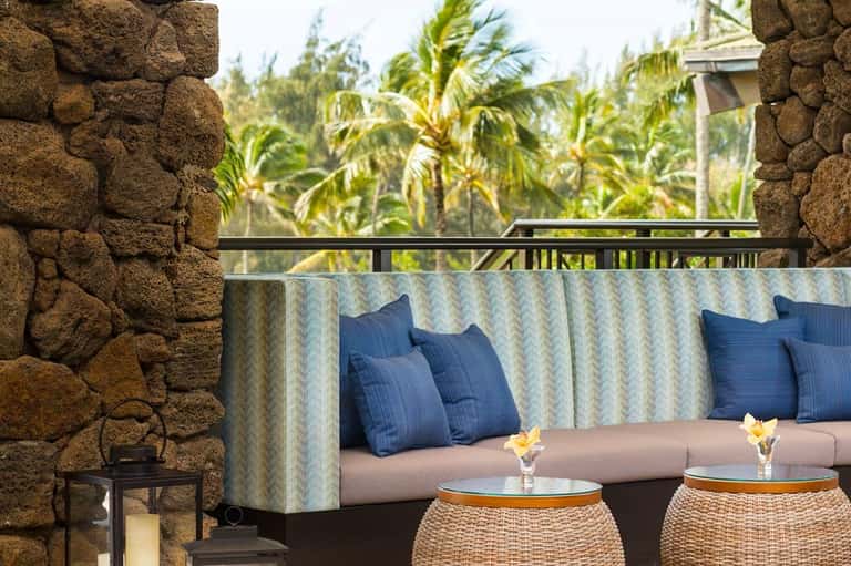 Outdoor lounge seating on stone terrace with cushioned sofa, blue pillows, tropical vegetation, and palm trees